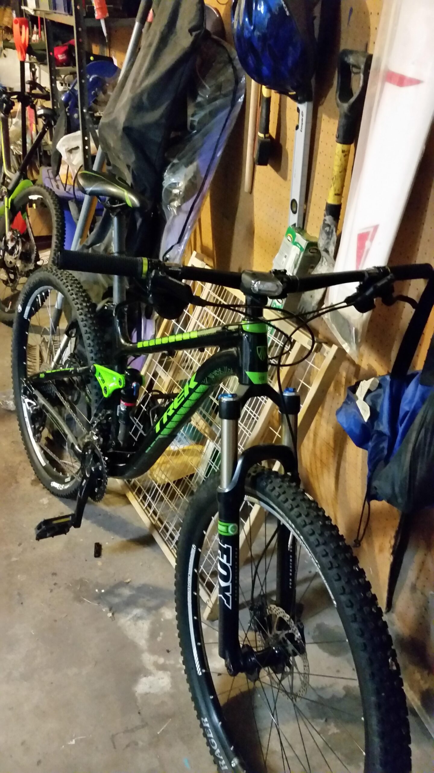 Trek Fuel EX 7 29er: A mountain bike with a black and green frame is positioned in a cluttered garage. Various tools and equipment, including a blue helmet and gardening tools, are visible in the background, along with a bicycle in the distance. The bike features thick tires and a suspension system, ready for outdoor adventures.