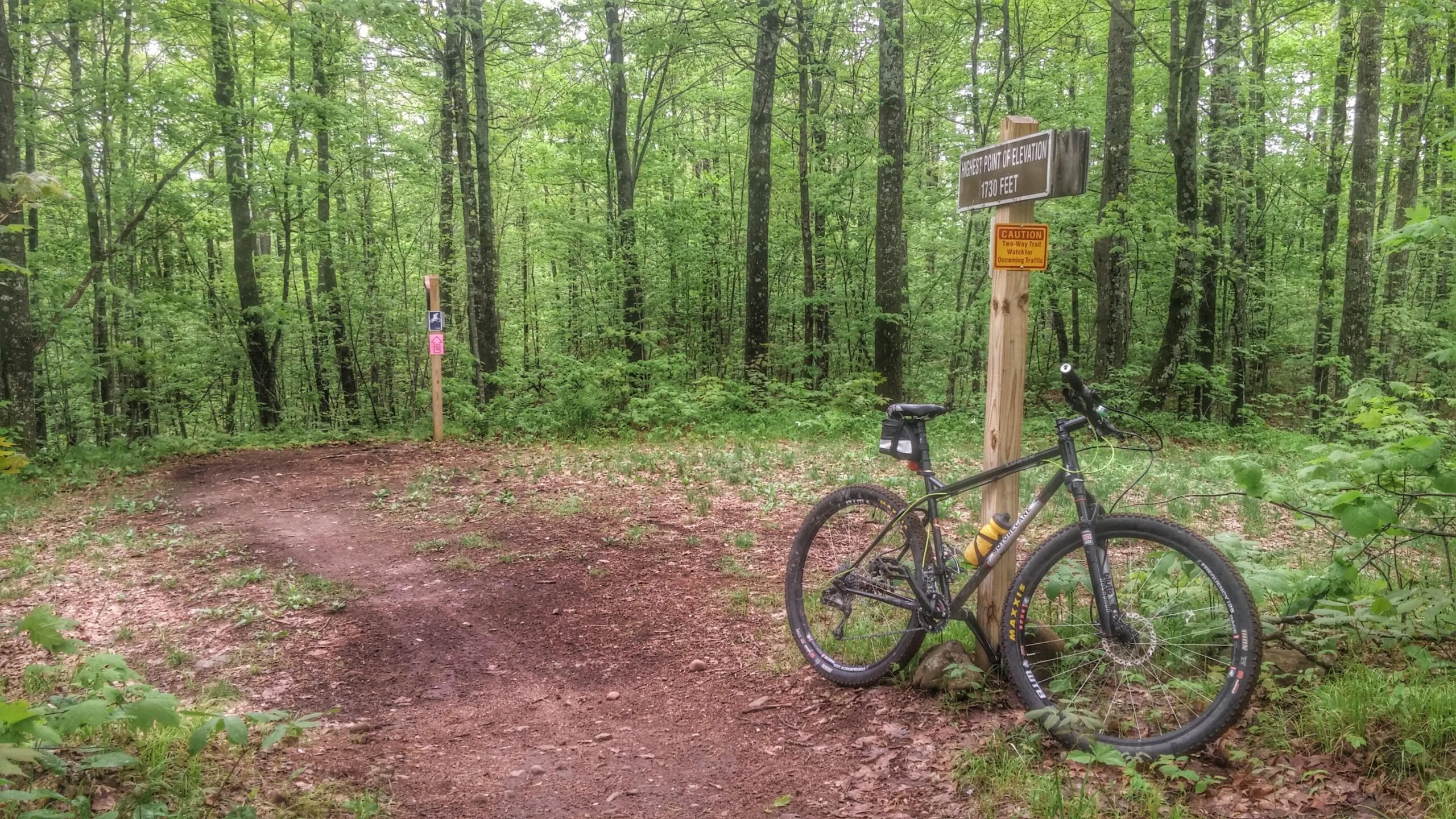 A mountain bike resting against a wooden sign indicating the "Highest Point of Elevation" at 1730 feet, set in a lush green forest with tall trees and vibrant foliage. A dirt path curves ahead, marked for hiking and biking trails, surrounded by a natural landscape. CAMBA: Hayward and Seeley Clusters mountain bike trail.