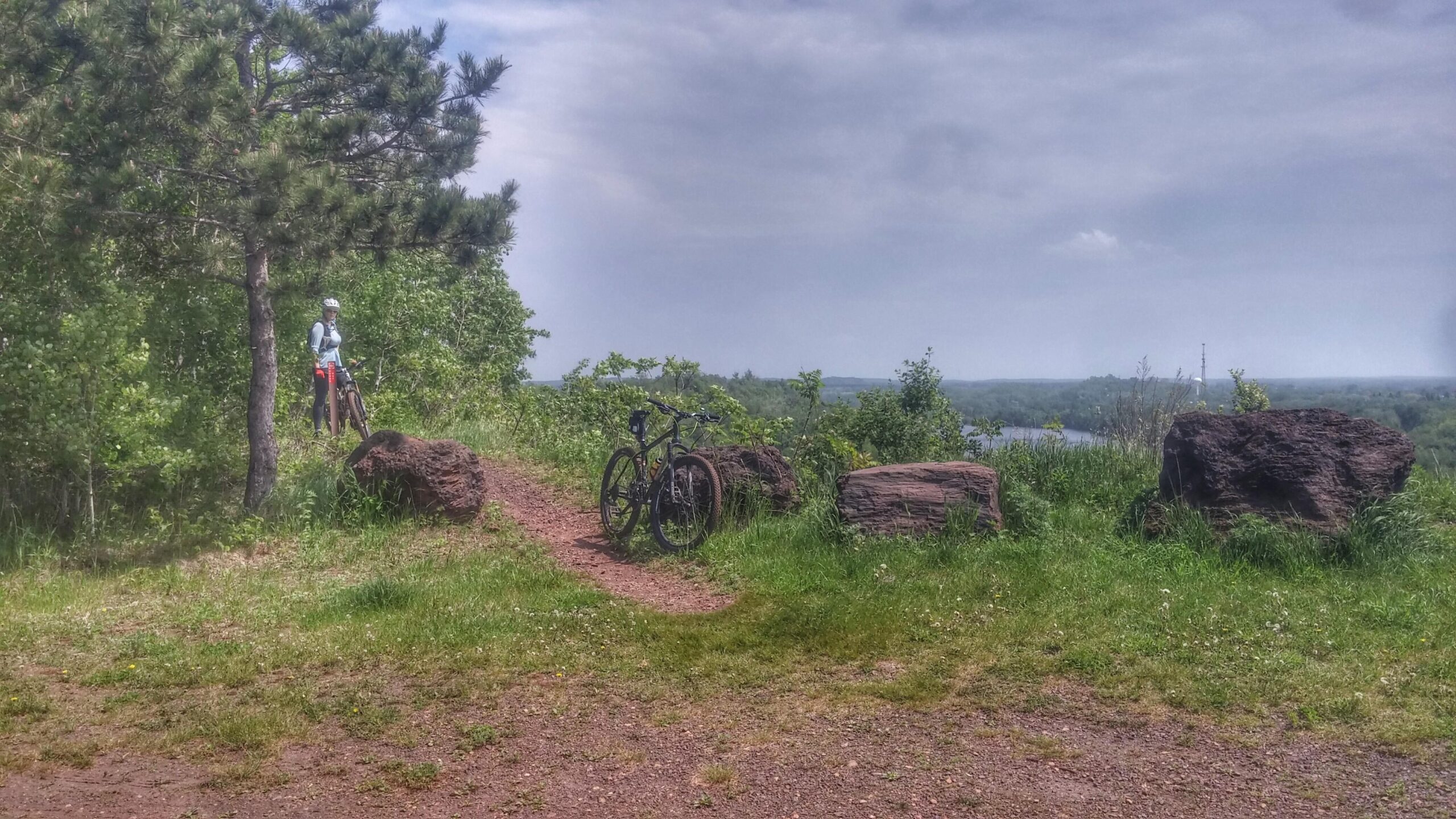 A scenic view from a trail, featuring a person standing beside their mountain bike near large rocks and surrounded by lush greenery. The background shows rolling hills and a river, under a partly cloudy sky. Cuyuna Lakes mountain bike trail.