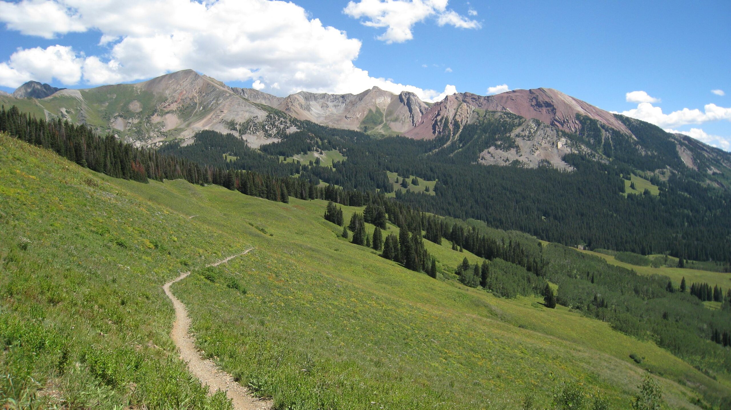 A scenic view of a mountainous landscape featuring green hills, a winding dirt path, and forested areas. The sky is bright blue with fluffy white clouds, and the rugged mountains in the background display various textures and colors. Trail 401 mountain bike trail.