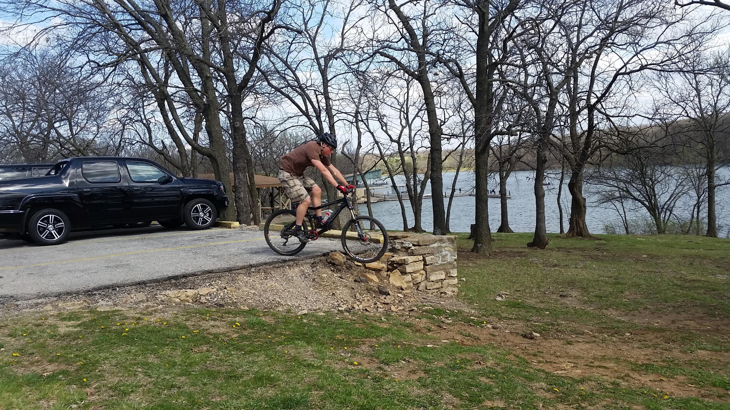 Trek Fuel EX 8: A mountain biker in shorts and a helmet hops off a small stone ledge onto a gravel path, with a lake and trees in the background. A black truck is parked nearby, and the scene is set in a natural outdoor environment.