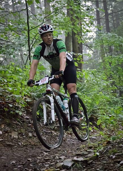 Trek Superfly AL Elite: A male mountain biker navigating a narrow, winding trail through a dense, green forest. He is wearing a green and black cycling jersey, black shorts, and a helmet, with a race number pinned to his jersey. The trail is surrounded by lush foliage and trees, creating a misty, serene atmosphere.