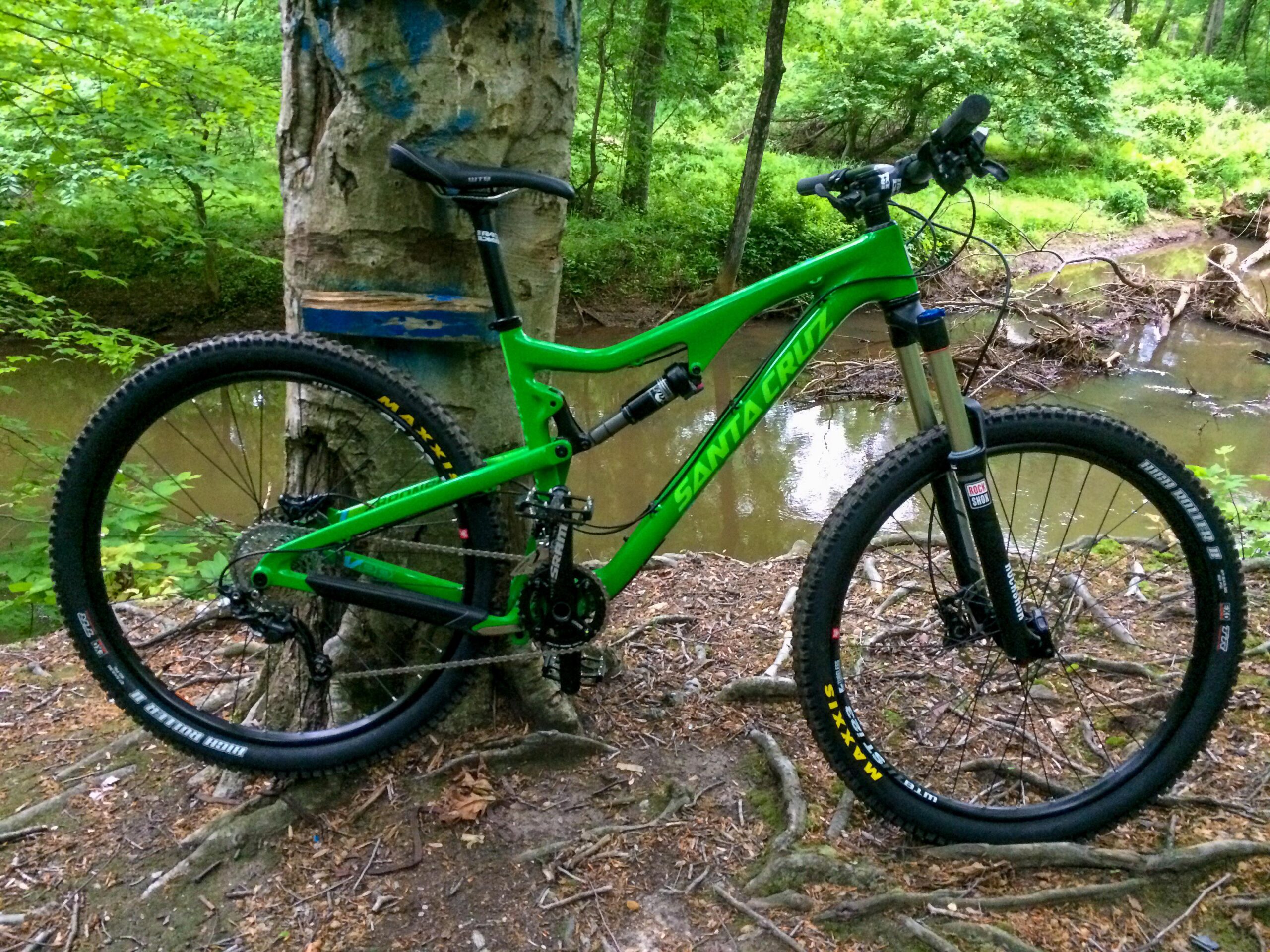 Santa Cruz Bronson: A vibrant green mountain bike is leaning against a tree trunk in a wooded area. The background features lush greenery and a creek, with visible roots and foliage on the ground. The bike’s wheels and components are detailed, showcasing its rugged design suitable for outdoor trails.