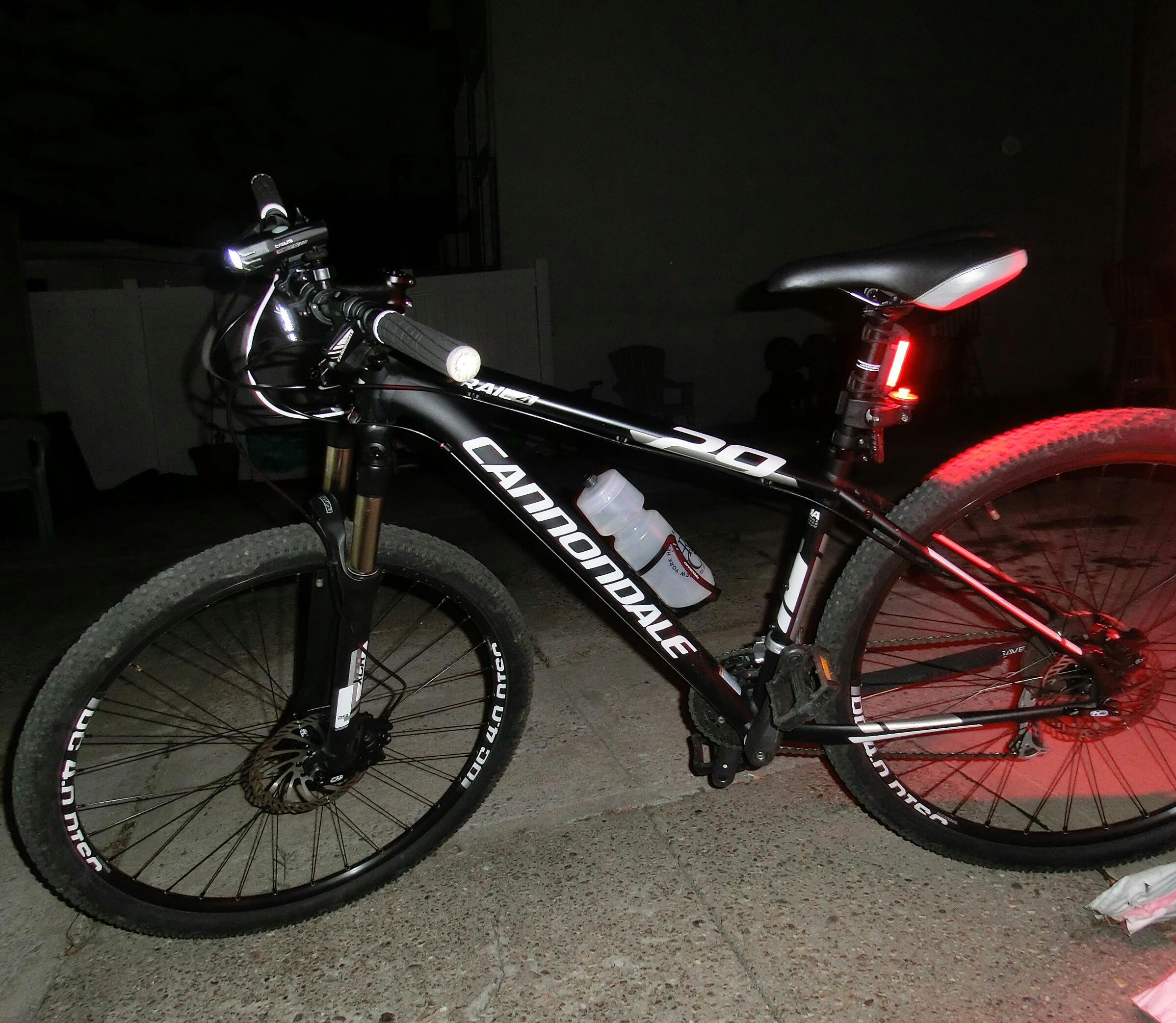 Cannondale Trail 29 4: A black Cannondale mountain bike parked on a concrete surface at night, featuring a front light, a rear red light, and a water bottle holder with a water bottle attached. The bike has knobby tires and is illuminated by a nearby light source.