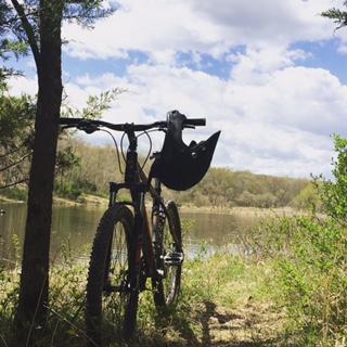 Giant Talon 27.5: A mountain bike resting against a tree, with a helmet hanging on the handlebars, by a serene lake surrounded by lush greenery and under a partly cloudy sky.