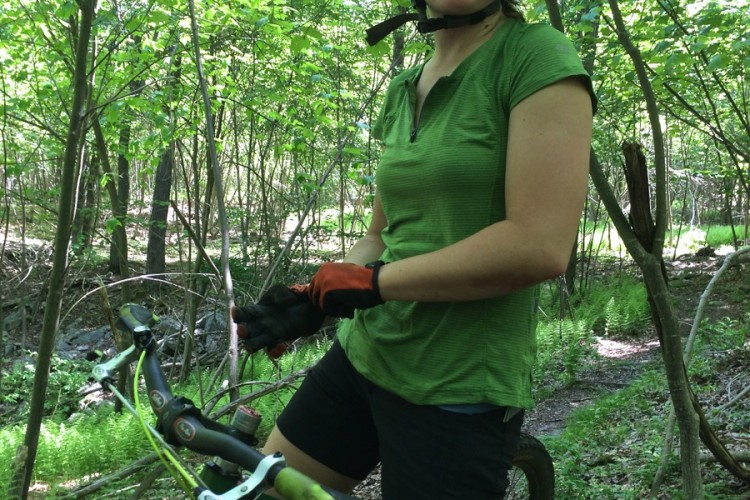 A person wearing a helmet and sunglasses stands beside a mountain bike in a lush green forest. They are dressed in a green shirt and black shorts, with their hands positioned on the bike's handlebars, appearing to be taking a break or preparing to ride. Sunlight filters through the leaves above, creating a vibrant natural setting.