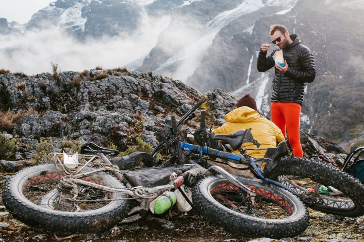 Two cyclists take a break in a mountainous area, surrounded by rocky terrain and misty peaks. One cyclist, wearing bright orange leggings and a black jacket, is eating from a package, while the other, dressed in a yellow jacket and brown beanie, is crouched beside their bikes. A couple of fat bikes, with muddy tires, lie on the ground nearby. The scene captures the essence of adventure and exploration in nature.