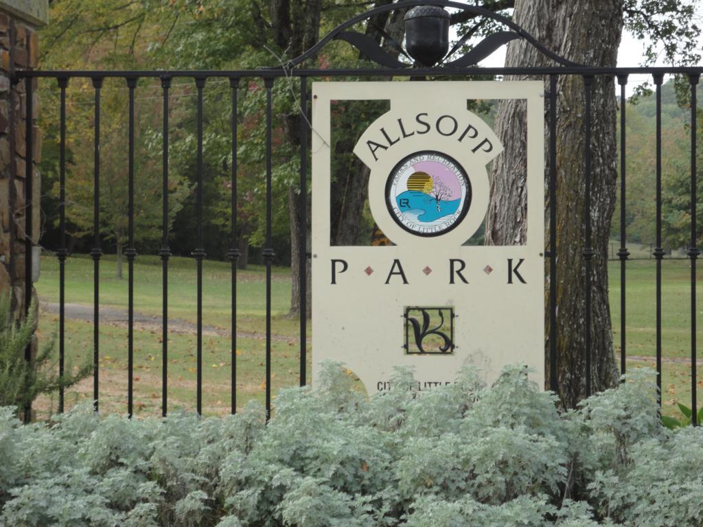 Signage for Allsopp Park, featuring a logo with a sun and tree, surrounded by greenery and a wrought iron gate. The park is set in a scenic area with trees in the background, indicating a natural environment. Allsopp Park mountain bike trail.