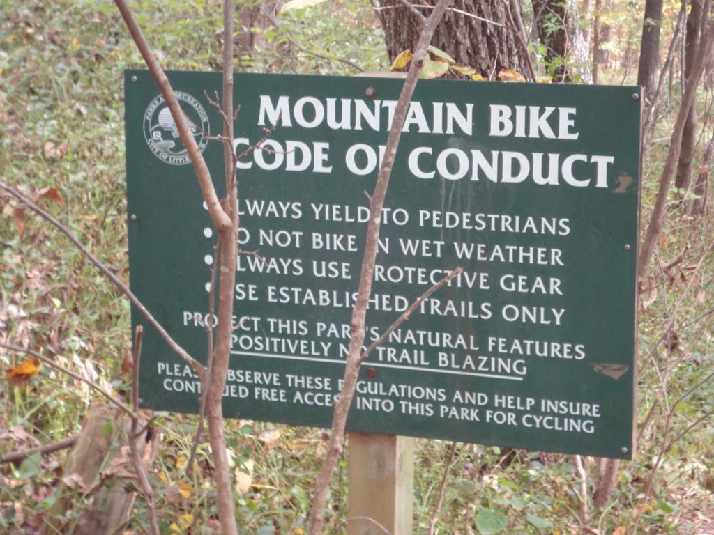 Green sign in a wooded area displaying the "Mountain Bike Code of Conduct," which includes guidelines such as yielding to pedestrians, avoiding biking in wet weather, using protective gear, and staying on established trails. The sign emphasizes protecting the park's natural features and encourages adherence to regulations. Allsopp Park mountain bike trail.