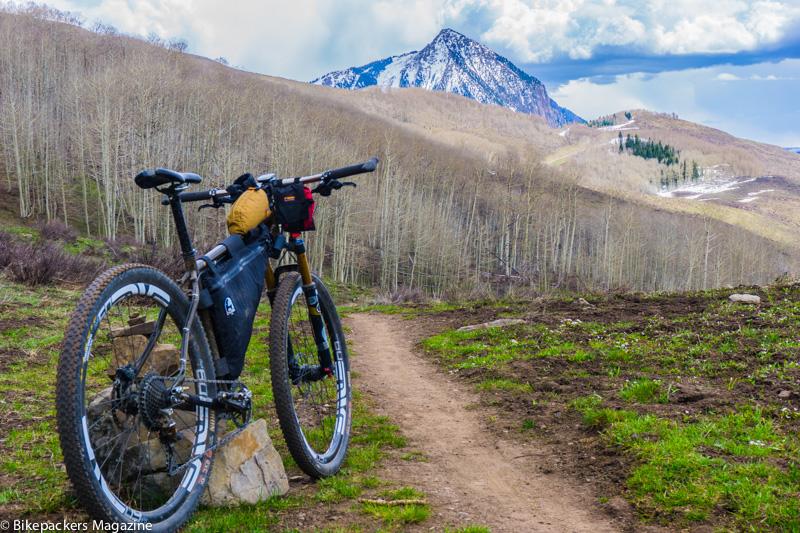 A mountain bike resting on a rock along a dirt trail, surrounded by golden aspens and green grass, with a backdrop of a snow-capped mountain and cloudy skies. Lupine Trail mountain bike trail.