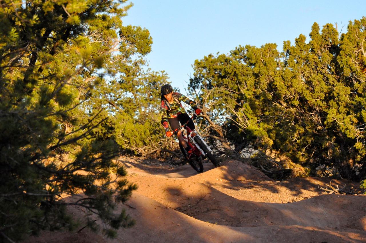 A mountain biker navigating a dirt trail surrounded by greenery and shrubs during golden hour, showcasing dynamic movement and skill on a gentle slope. Hustle and Flow mountain bike trail.