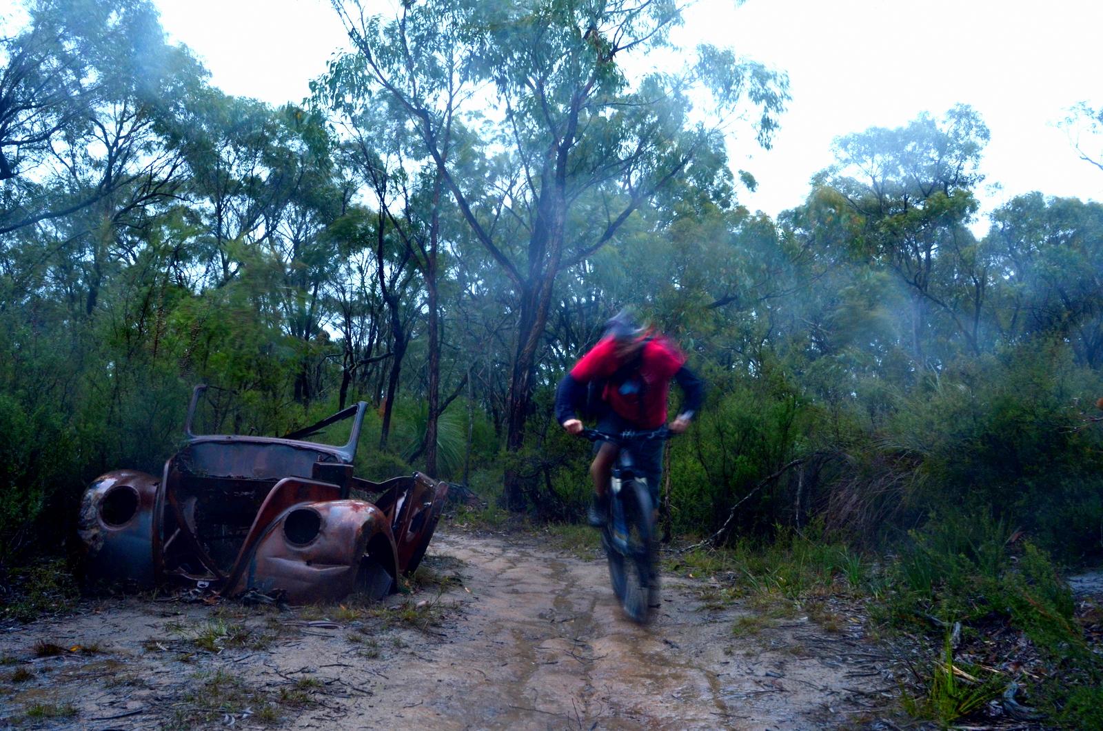 A cyclist rides along a muddy trail in a forested area, with an old, rusted car frame visible to the side. The scene is misty and rainy, characterized by lush greenery and tall trees. Forrest Mtb Trails mountain bike trail.