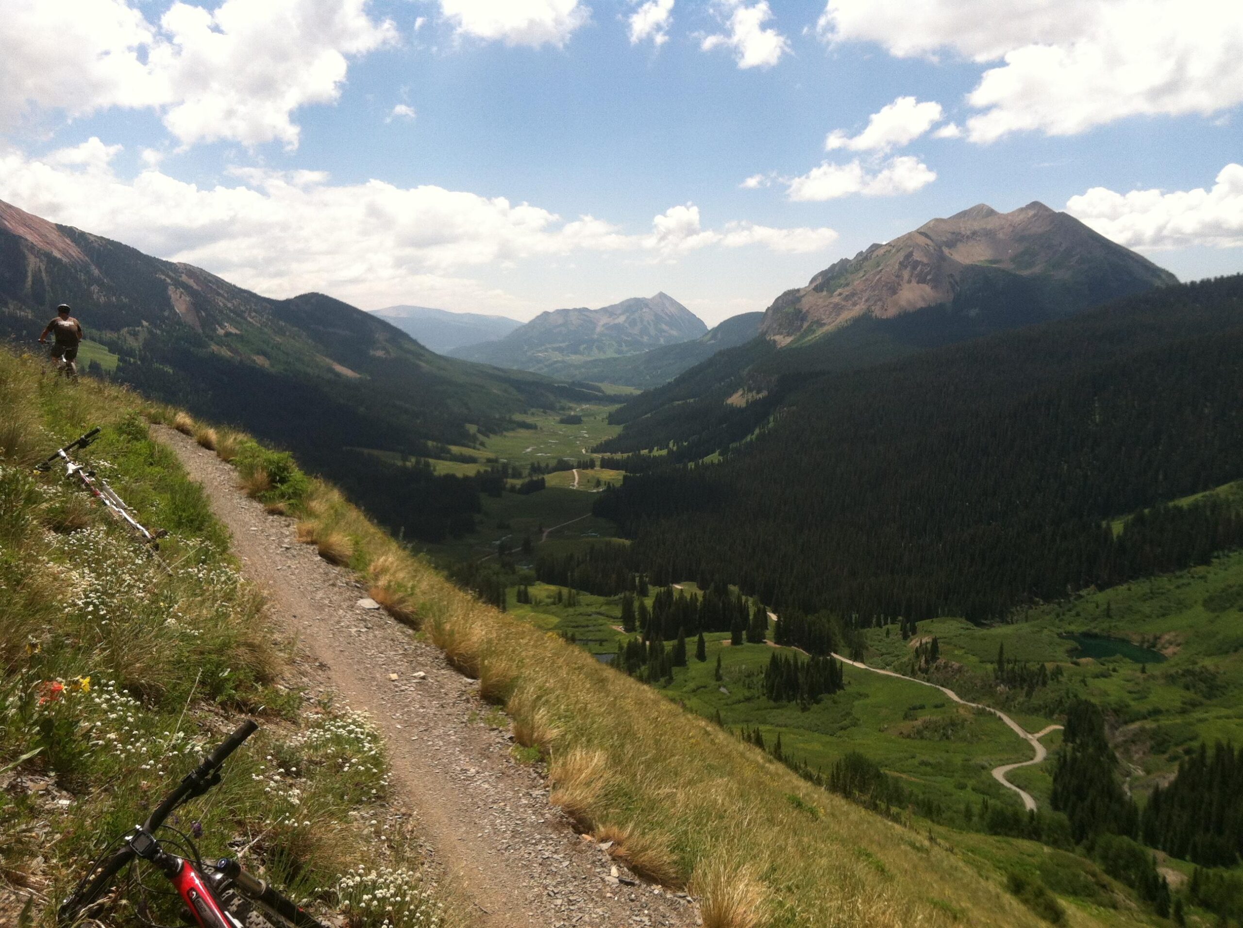 A scenic view from a mountain biking trail overlooking a lush green valley surrounded by mountains. A cyclist stands on the trail, which is lined with tall grass and wildflowers, while mountain bikes are resting nearby. The sky is partly cloudy, adding to the picturesque landscape. Trail 401 mountain bike trail.