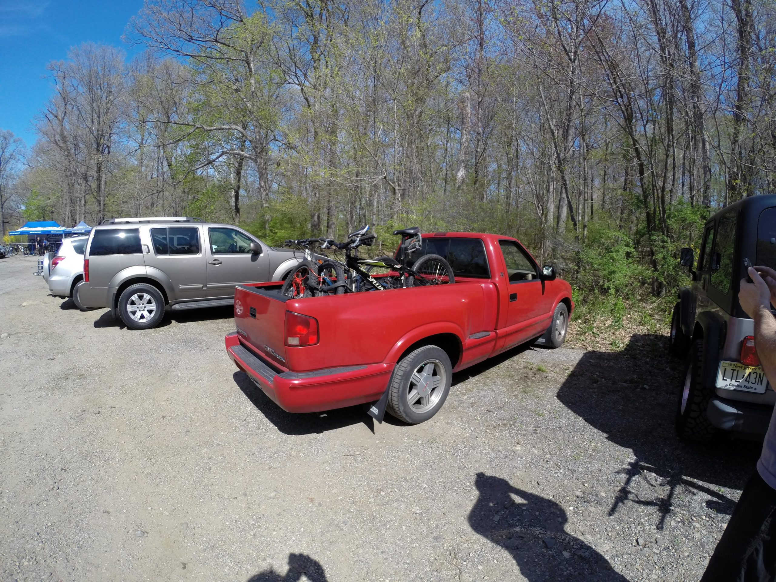 A red pickup truck parked in a gravel area, with two bicycles secured in the truck bed. In the background, several other vehicles are visible, along with sparse trees and a clear blue sky. The image is set in a natural outdoor environment. Ringwood Skylands Manor mountain bike trail.
