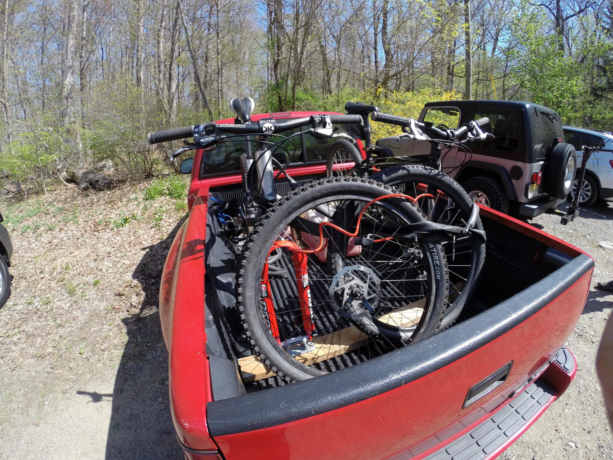 Alt text: A red pickup truck parked in a wooded area, with two mountain bikes securely loaded in the truck bed. The scene is sunny with leafless trees and green foliage in the background. Ringwood Skylands Manor mountain bike trail.