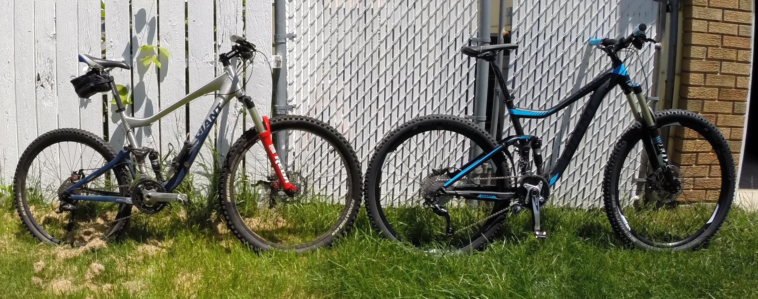 Giant Trance 27.5 2: Two mountain bikes parked on grass beside a white wooden fence. The left bike features a silver and blue frame with a red fork, while the right bike has a black frame with blue accents. Both bikes have thick, durable tires suited for off-road riding.