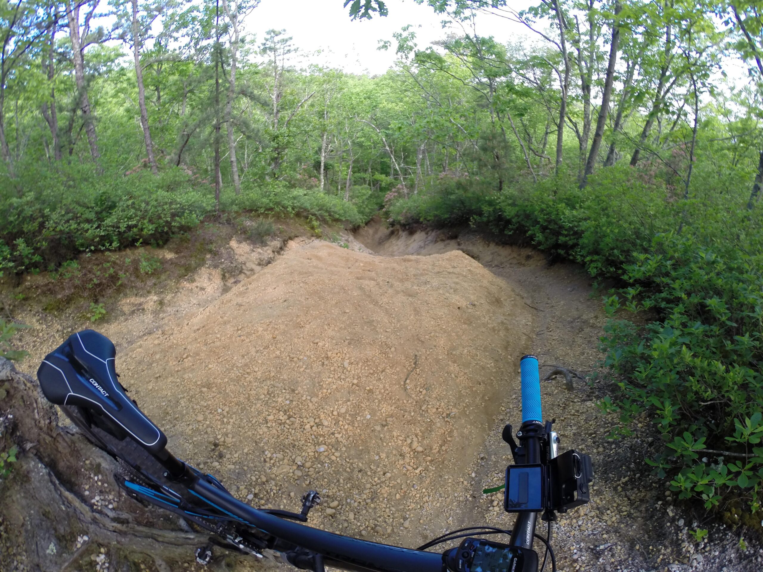 Image depicts a mountain bike handlebar with a blue grip, camera mount, and a bike saddle in the foreground. In the background, a dirt path leads into a wooded area, surrounded by greenery and trees. The ground is uneven and there is a pile of dirt along the path. Allaire State Park mountain bike trail.