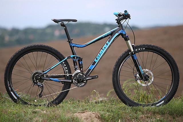 Giant Stance 27.5 2: A blue and black mountain bike is positioned on grassy terrain, with a blurred landscape in the background. The bike features thick tires, a lightweight frame, and visible components like the handlebars and gears.