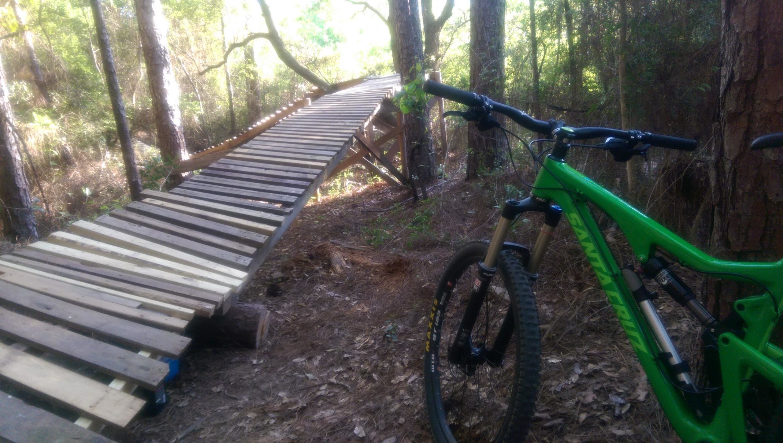A mountain bike resting on the ground beside a wooden bridge in a forested area. The bridge is constructed from wooden planks and is elevated off the ground, surrounded by trees and shrubbery. UWF Mountain Bike Trails mountain bike trail.