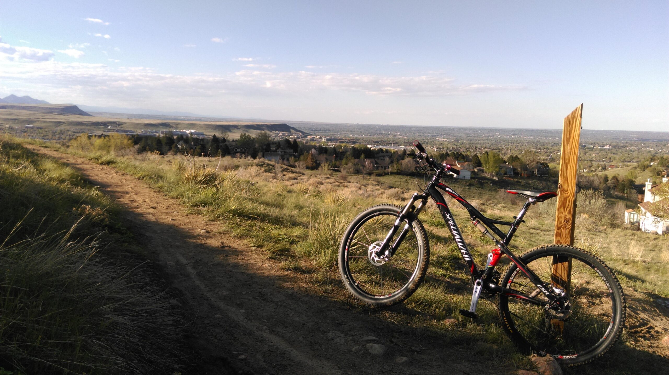 Specialized Epic Comp: A mountain bike parked on a dirt trail overlooking a scenic landscape. In the background, rolling hills and a valley can be seen under a partly cloudy sky. The foreground features tall grasses and greenery along the path.