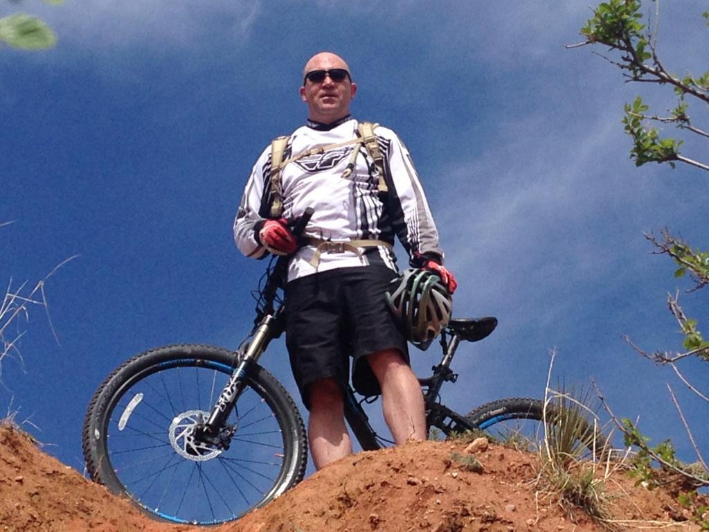 An athlete in cycling gear stands confidently on a hilltop with a mountain bike beside him. He is wearing sunglasses, a protective helmet, gloves, and a jersey, surrounded by blue sky and sparse vegetation. The scene captures a moment of accomplishment in a cycling adventure. Red Rock Canyon mountain bike trail.