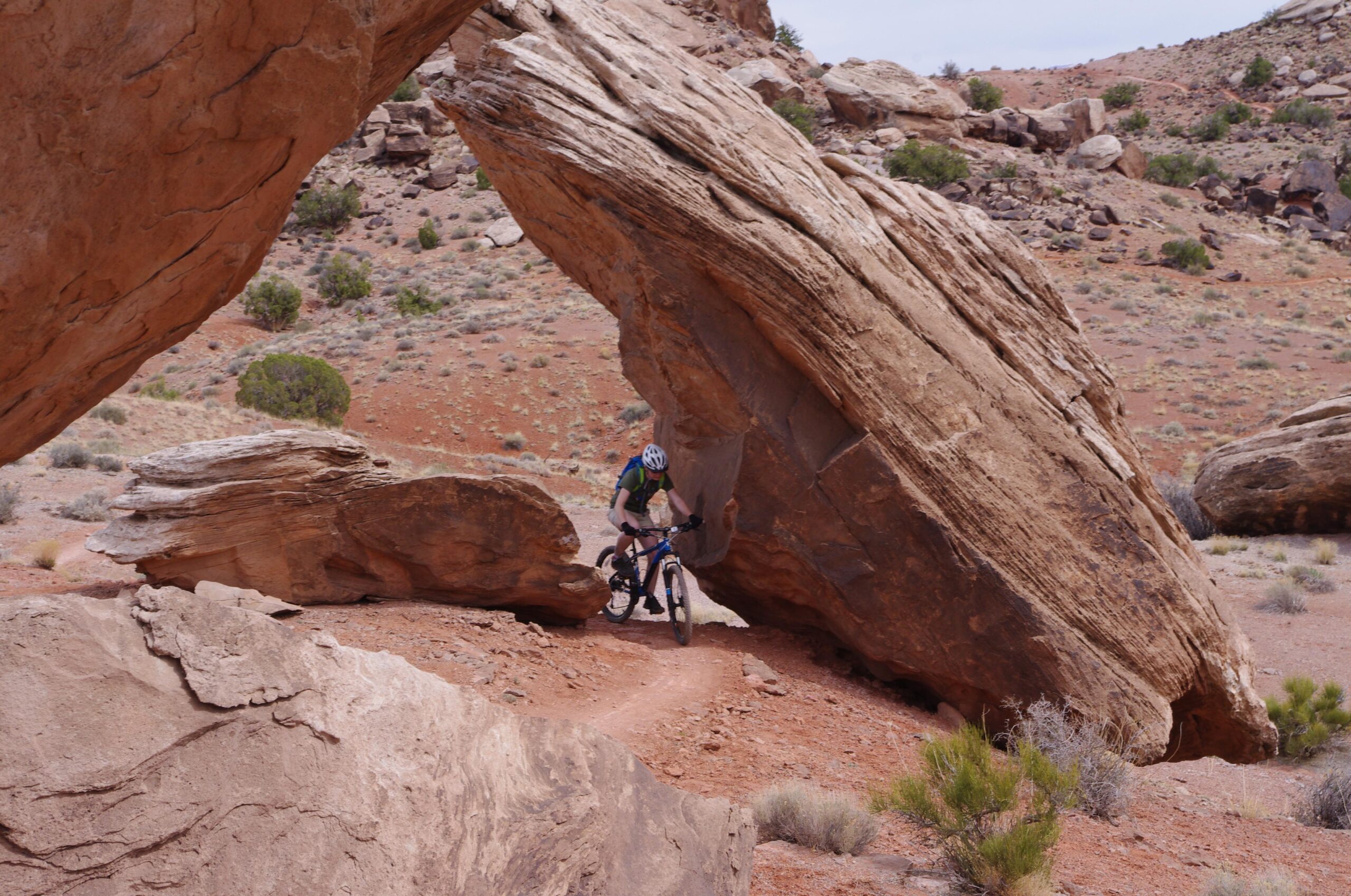 A mountain biker navigating through a narrow passage between two large rock formations in a desert landscape. The path is dirt, with reddish soil and sparse vegetation in the background. Moab Brand Trails mountain bike trail.