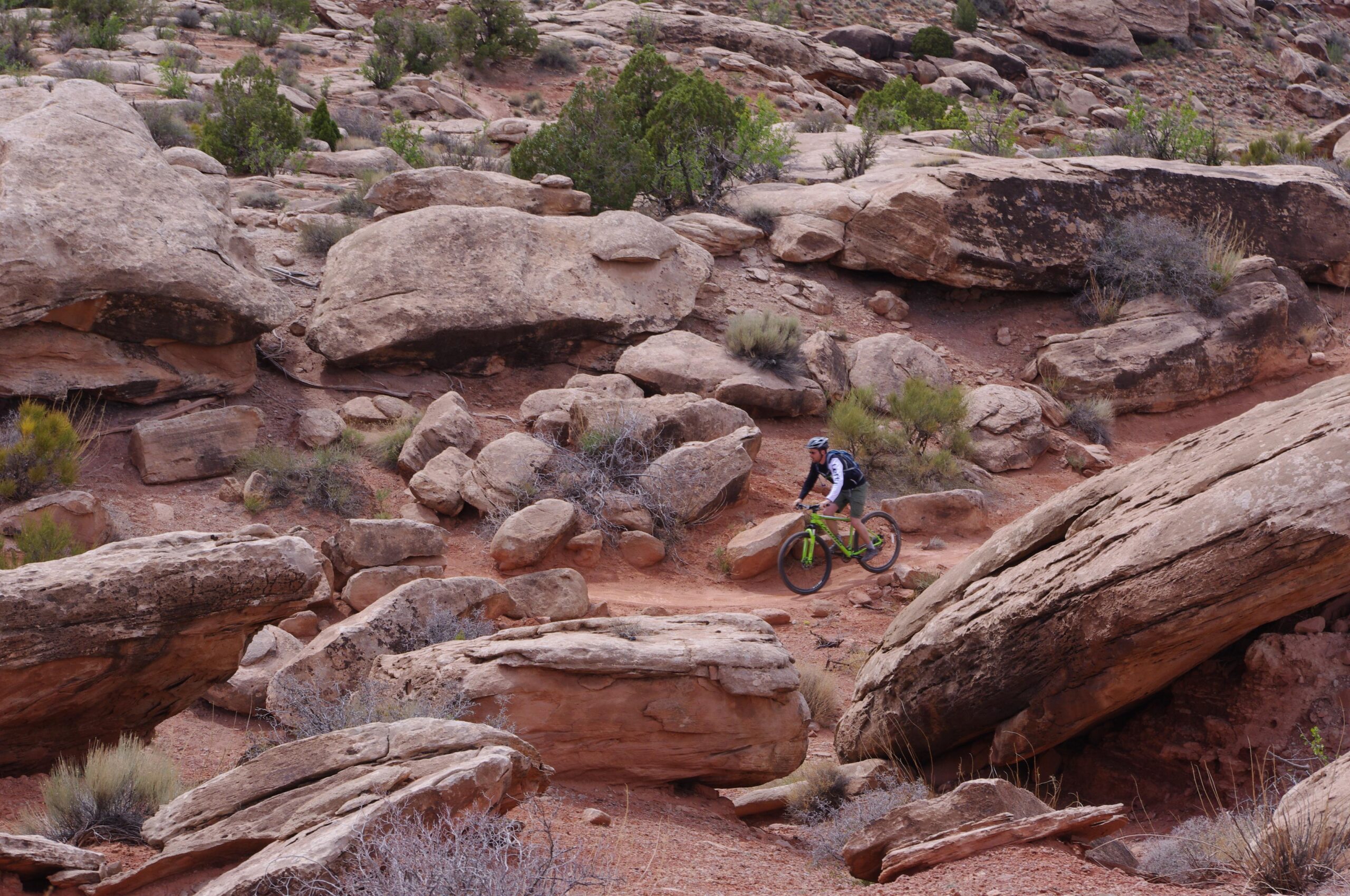 A mountain biker navigating a rocky path surrounded by large boulders and sparse vegetation in a rugged outdoor landscape. Moab Brand Trails mountain bike trail.