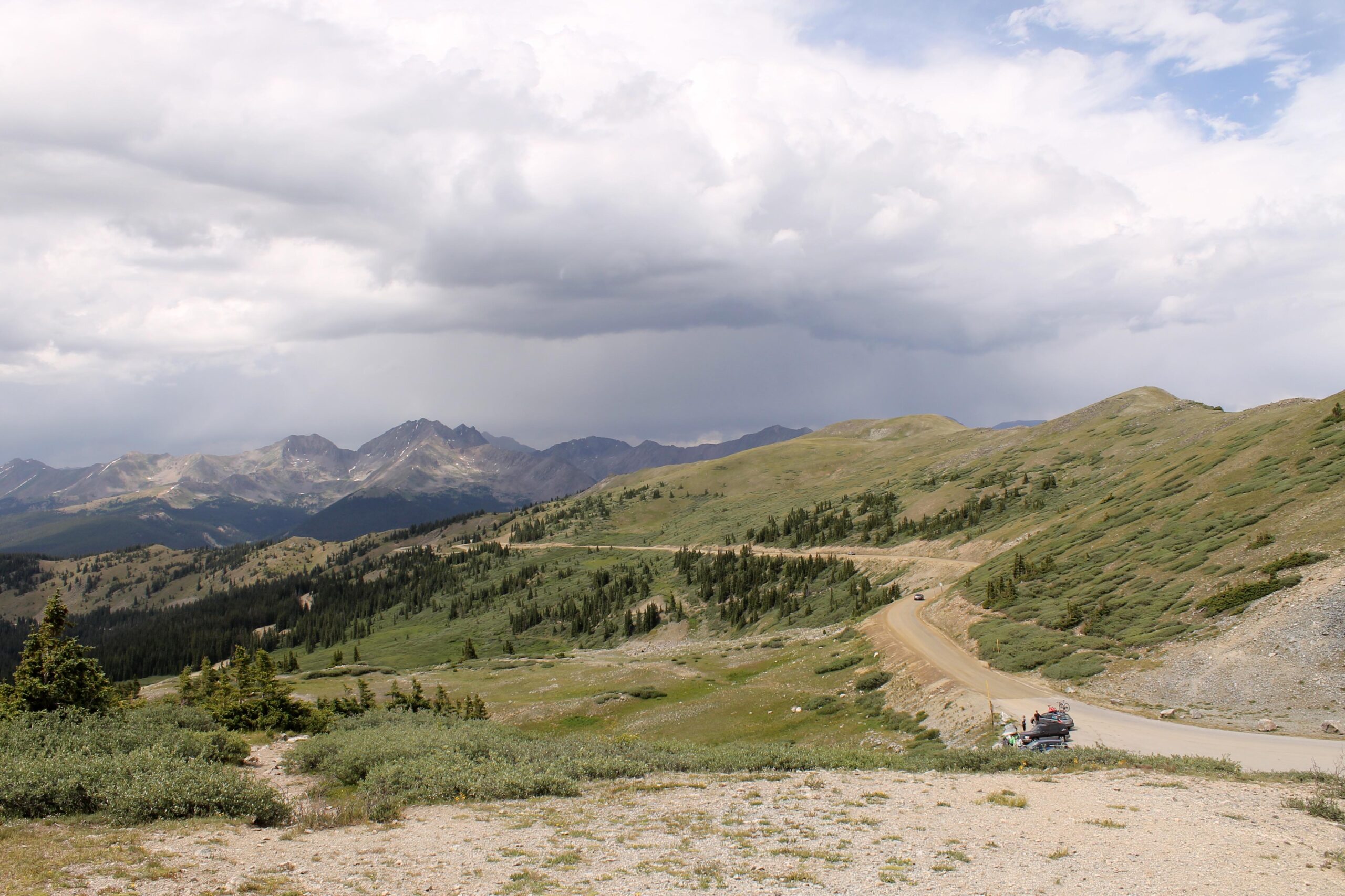 A scenic view of mountainous terrain under a cloudy sky, featuring rolling green hills, trees, and a winding dirt road. A parked vehicle is visible on the roadside, suggesting a peaceful outdoor setting for exploration. Cottonwood Pass Road mountain bike trail.