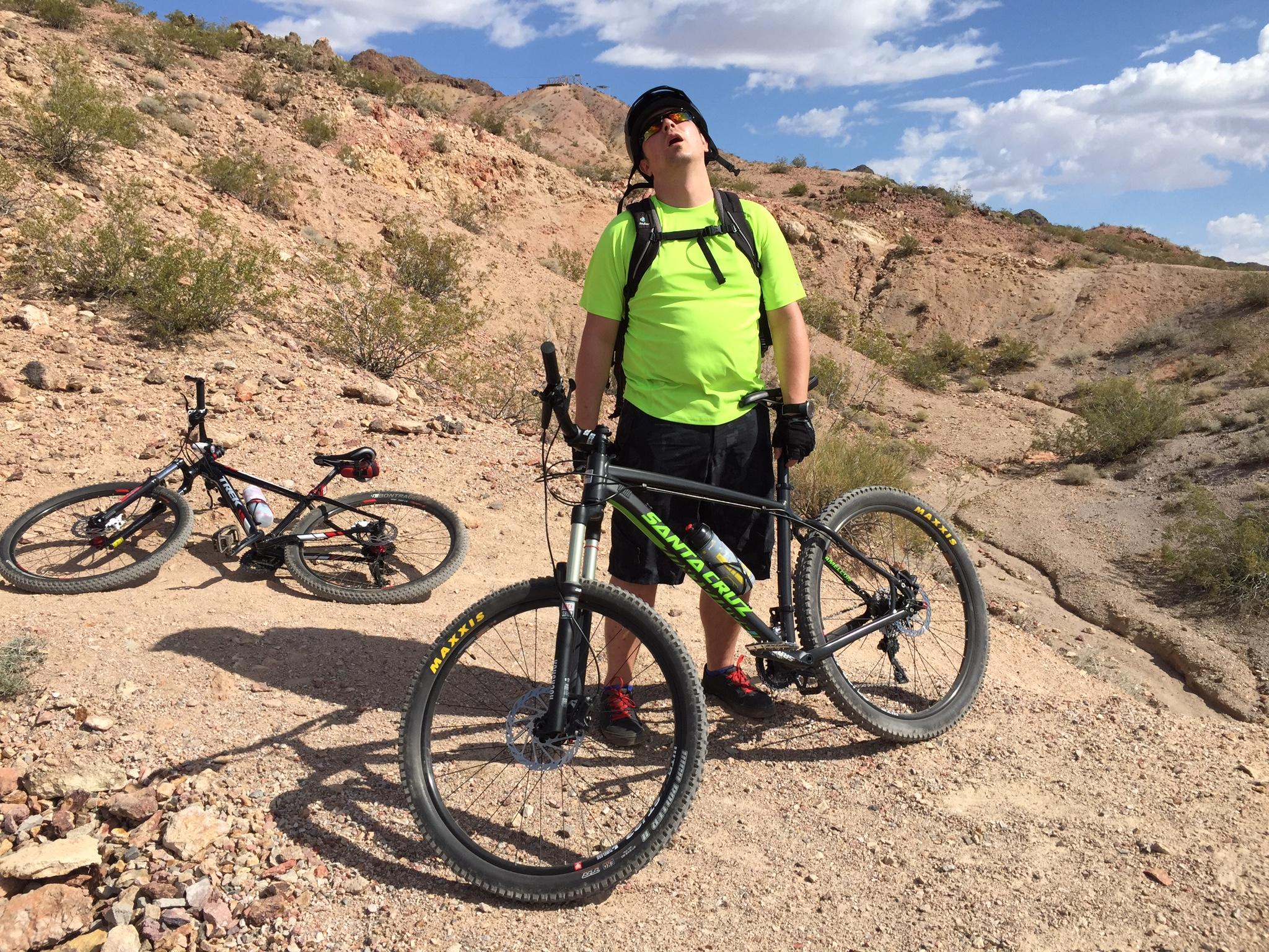 A person wearing a bright green shirt and a helmet stands next to a mountain bike on a rocky trail, appearing to take a break. A second bike lies on the ground nearby. The landscape features dry, hilly terrain with sparse vegetation and a blue sky with scattered clouds. Bootleg Canyon mountain bike trail.