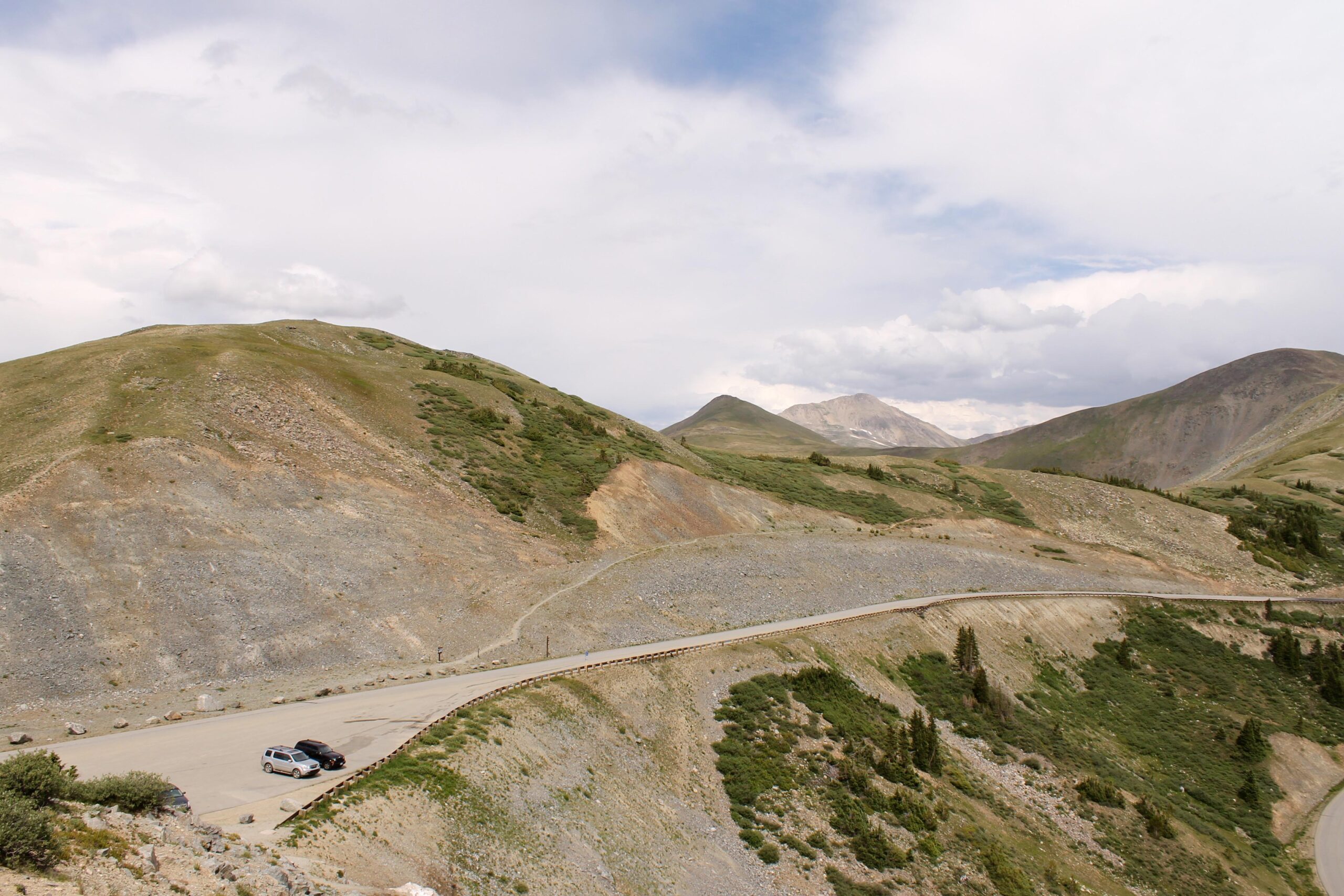 A scenic view of a winding road that curves around a mountainous landscape, featuring green hills and rocky terrain under a cloudy sky. Two parked vehicles are visible on the roadside, with mountains in the background. Cottonwood Pass Road mountain bike trail.