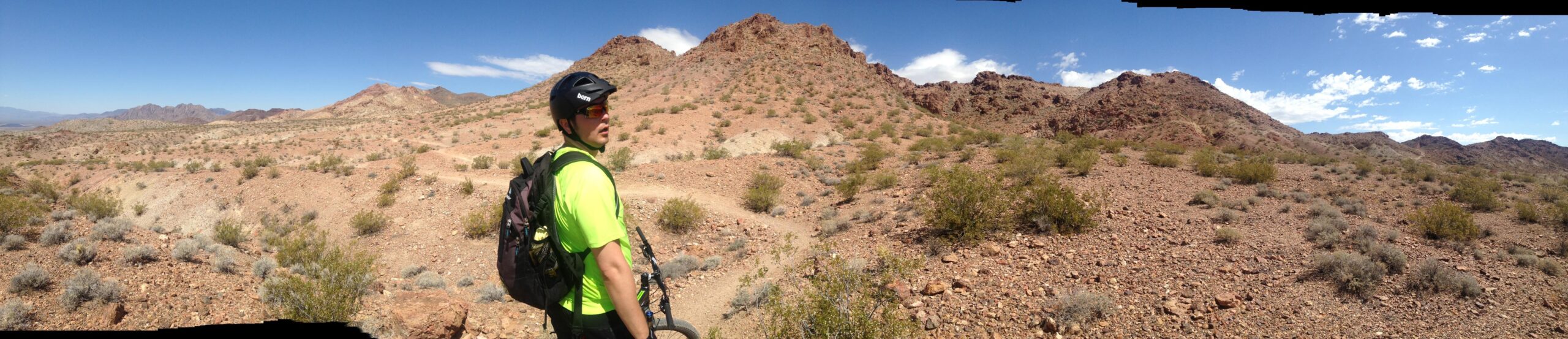 A person wearing a bright green shirt, helmet, and backpack stands on a rocky trail in a desert landscape, surrounded by mountainous terrain and sparse vegetation under a clear blue sky. Bootleg Canyon mountain bike trail.