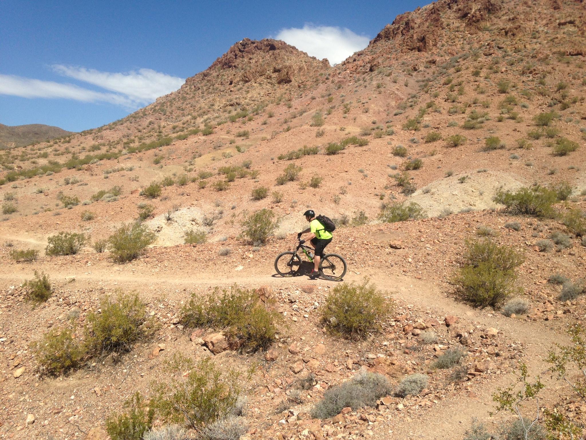A person riding a mountain bike on a dirt trail in a rocky, arid landscape, surrounded by small shrubs and hills under a clear blue sky. Bootleg Canyon mountain bike trail.