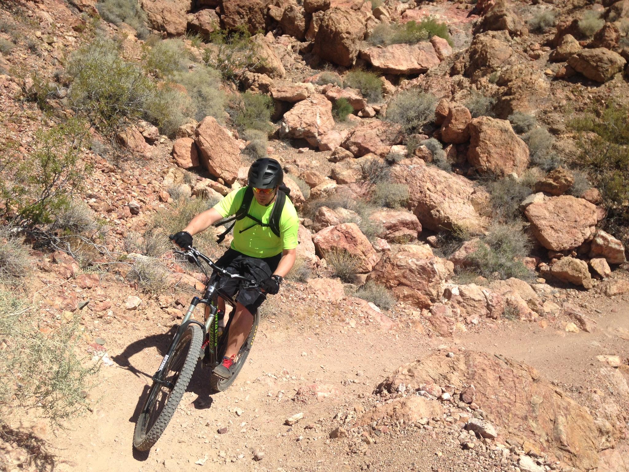 A mountain biker wearing a bright yellow shirt and helmet is riding on a rocky trail surrounded by dry shrubs and boulders. The sun is shining down on the rugged terrain, highlighting the challenging landscape. Bootleg Canyon mountain bike trail.
