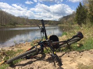 Giant Talon 27.5: A mountain bike resting on the ground near a calm river, surrounded by greenery and trees. The sky is partly cloudy, suggesting a pleasant outdoor atmosphere.