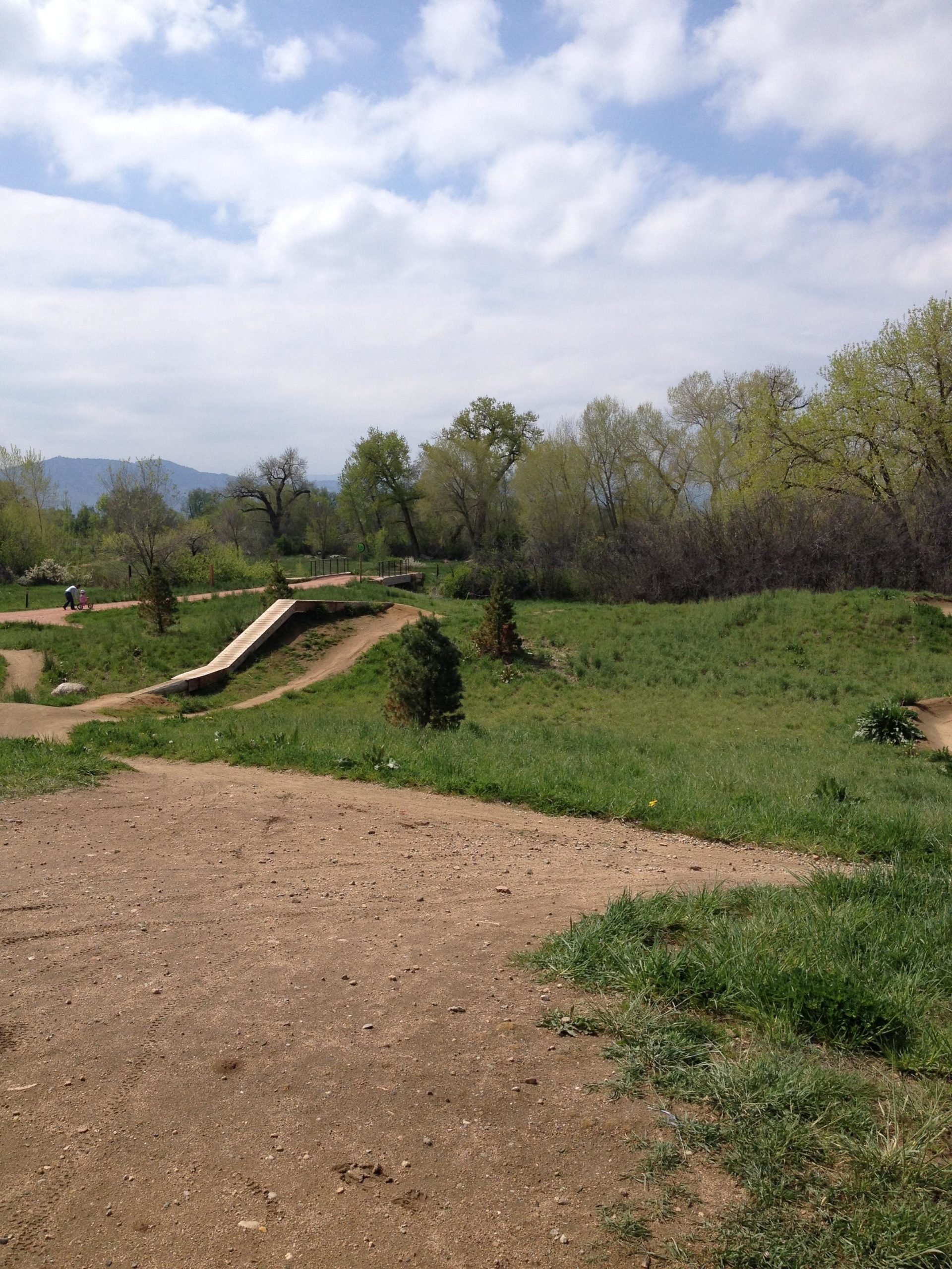 A scenic outdoor area featuring a dirt path winding through greenery, with small jumps and ramps for biking. The background includes trees and distant hills under a partly cloudy sky. A person is visible working on the path in the foreground. Valmont Bike Park mountain bike trail.
