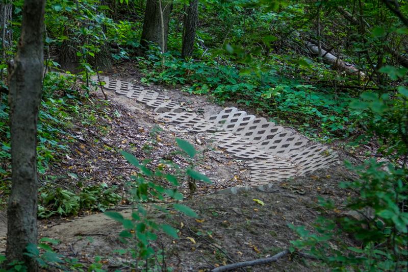 A winding dirt pathway in a wooded area, featuring a series of textured, patterned matting laid along the trail to provide stability and traction. Surrounding vegetation includes green leaves and underbrush, creating a natural setting. Illiniwek Forest Preserve mountain bike trail.