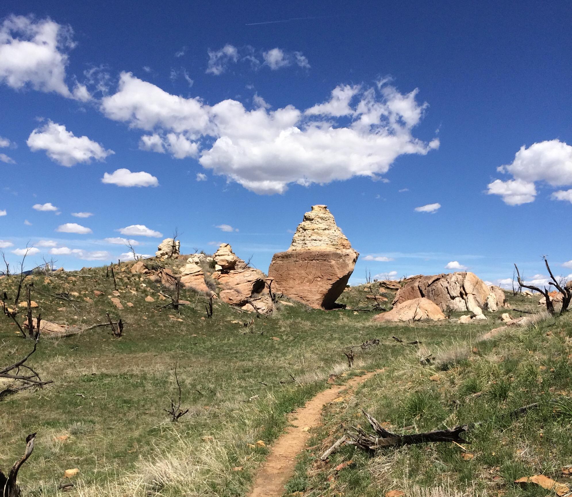 A rocky landscape featuring distinct rock formations under a blue sky with fluffy clouds. A dirt path winds through green grass and sparse vegetation, hinting at a natural setting that includes some burned tree remnants. Oil Well Flats mountain bike trail.