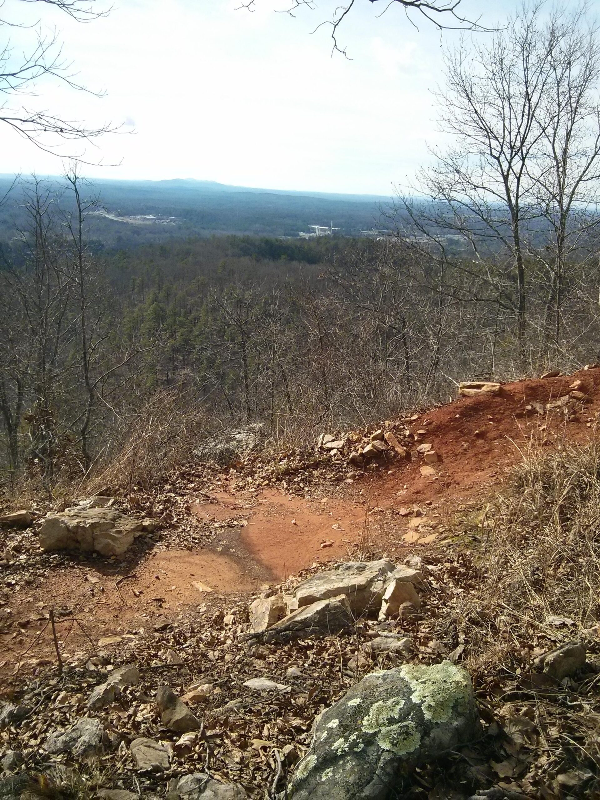 A panoramic view from a rocky outcrop overlooking a lush, wooded valley. The landscape features rolling hills in the distance, with a mix of bare trees and green foliage. The ground is covered with red earth and scattered rocks, while fallen leaves add texture to the scene. The sky is partly cloudy, contributing to the serene atmosphere of the natural setting. Coldwater Mountain mountain bike trail.