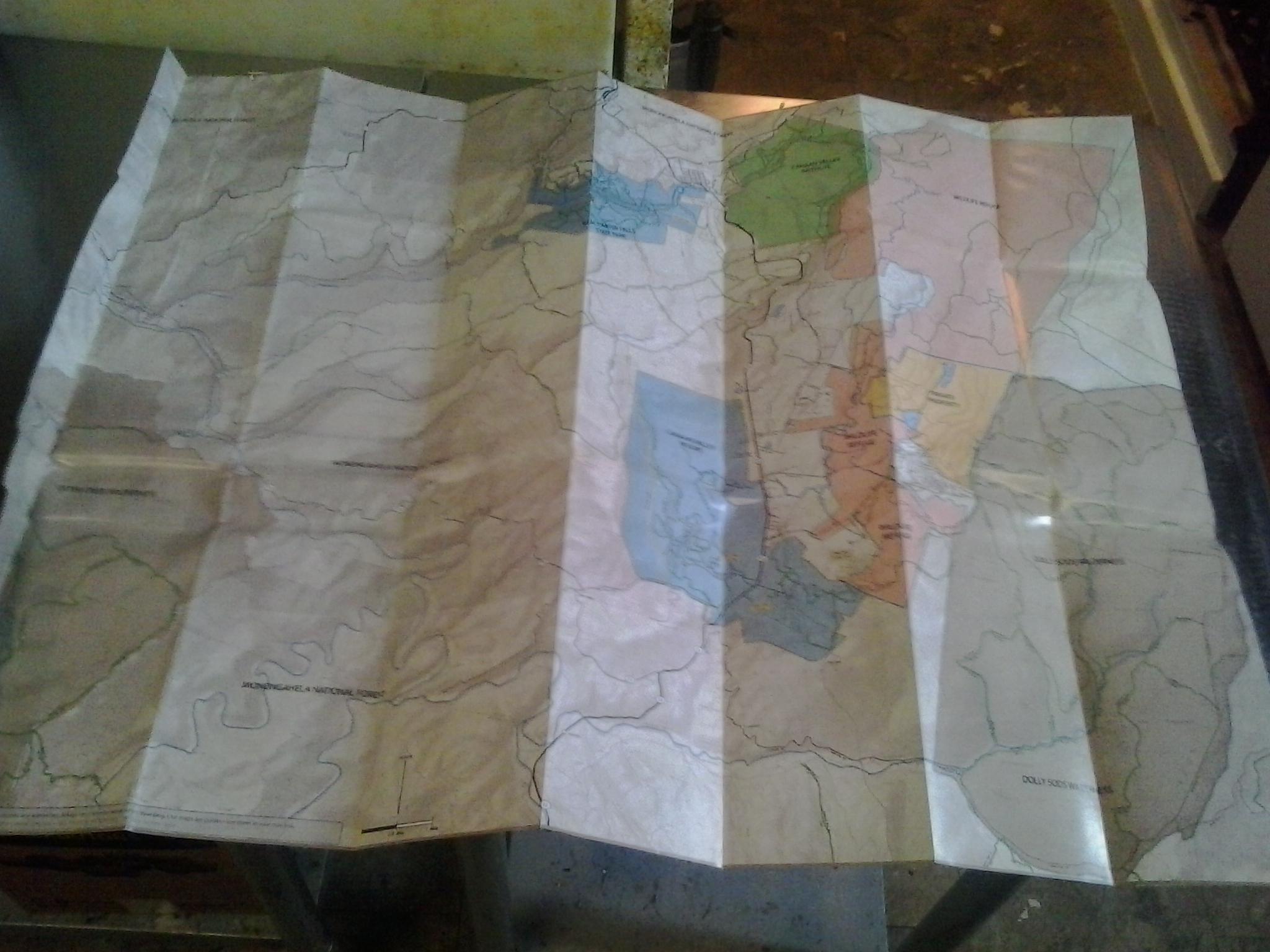 Cannan Valley Explorer Trail Map: A folded topographic map displaying various colorful sections indicating different areas, with contour lines illustrating the terrain. The map includes labels for features such as national forests and parks. Light reflects off the paper, highlighting its creases and textures.