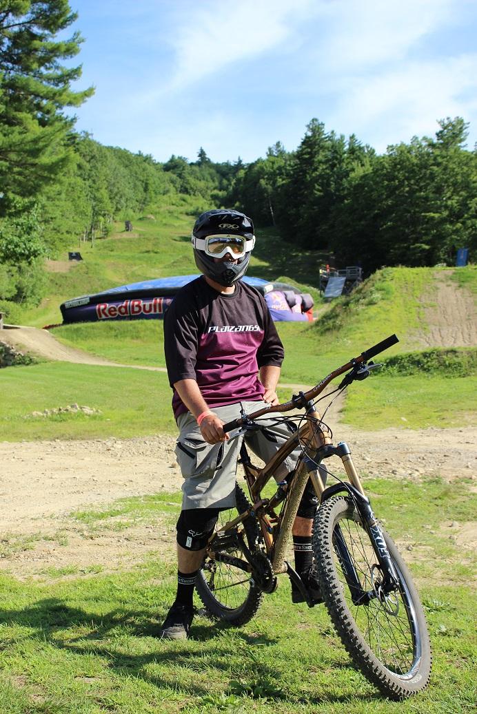 Transition Covert: A mountain biker wearing a helmet and protective gear stands next to a bicycle on a grassy area with a dirt track in the background. The track features hills and a large inflatable obstacle with a Red Bull logo.