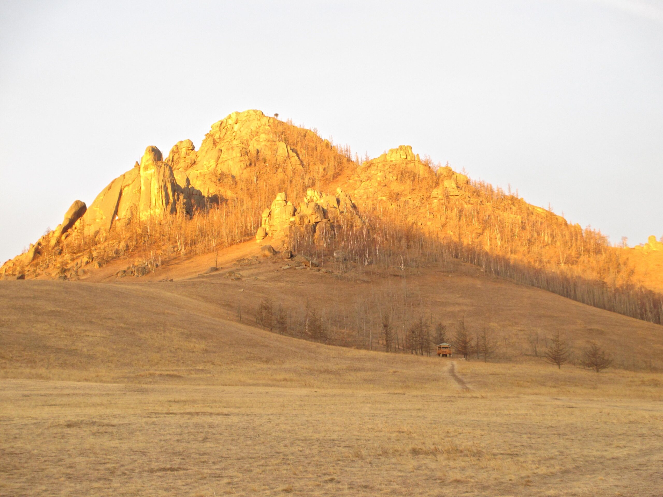 A scenic view of a mountainous landscape during sunset, featuring a golden-hued rocky hill with sparse trees. A pathway leads towards a small wooden structure at the base of the hill, with dry grass covering the foreground. The sky is clear, creating a serene atmosphere. Camel Run mountain bike trail.