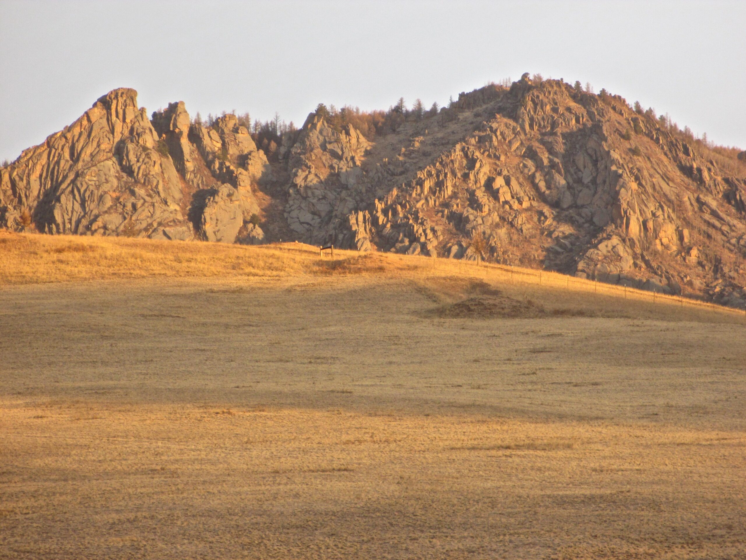 A rugged landscape featuring prominent rock formations atop a grassy hillside, illuminated by warm evening light. The foreground consists of a plain with sparse vegetation, leading up to the dramatic, jagged mountains in the background. Camel Run mountain bike trail.