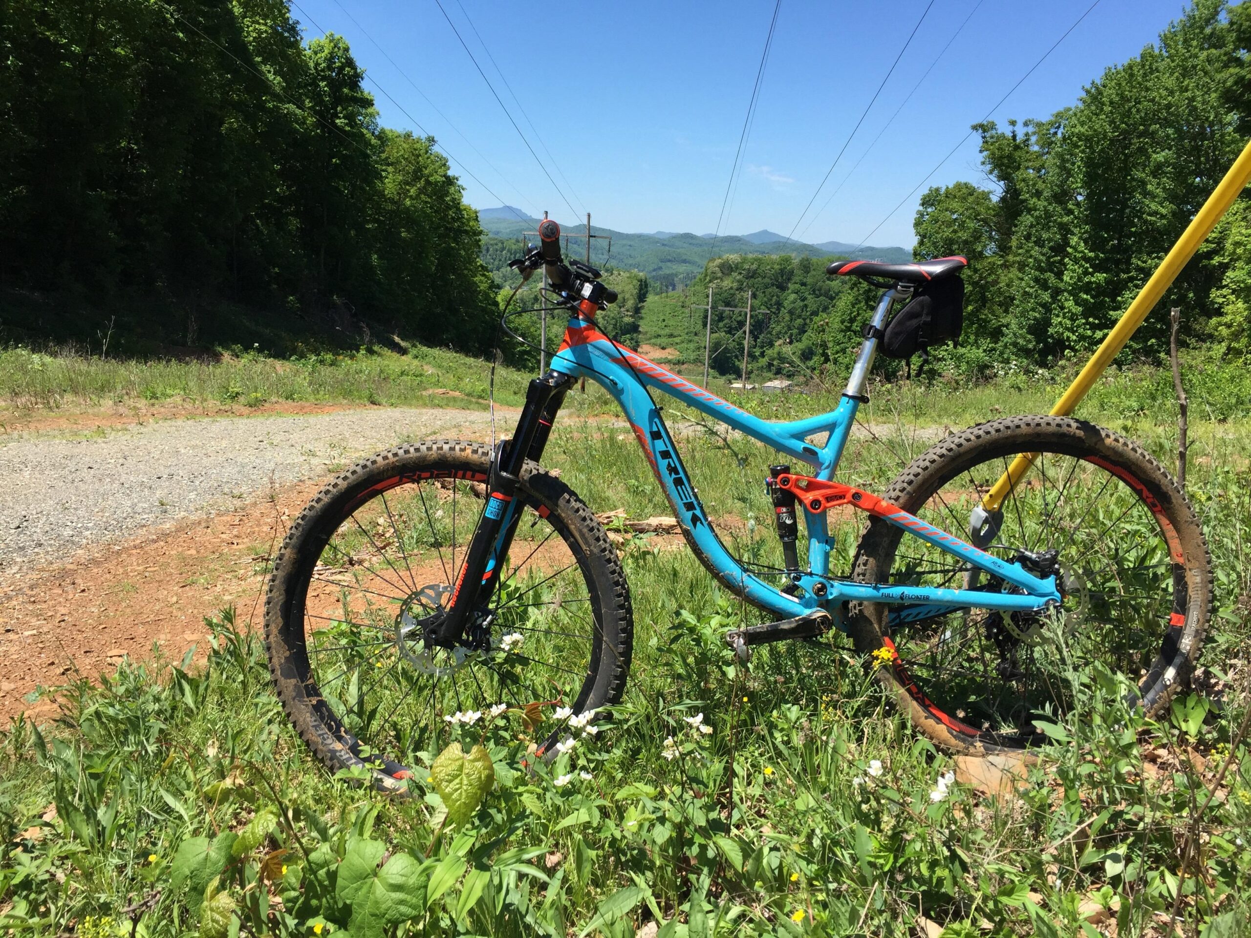 A brightly colored mountain bike resting on a gravel path surrounded by green grass and wildflowers, with trees and distant mountains in the background under a clear blue sky. Rocky Knob Park mountain bike trail.