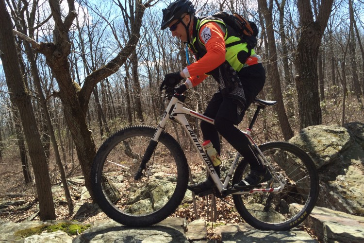 A mountain biker in a bright orange and green outfit skillfully navigating over rocky terrain in a forested area. The cyclist is wearing a helmet and riding a white mountain bike, with a backdrop of bare trees and blue sky.