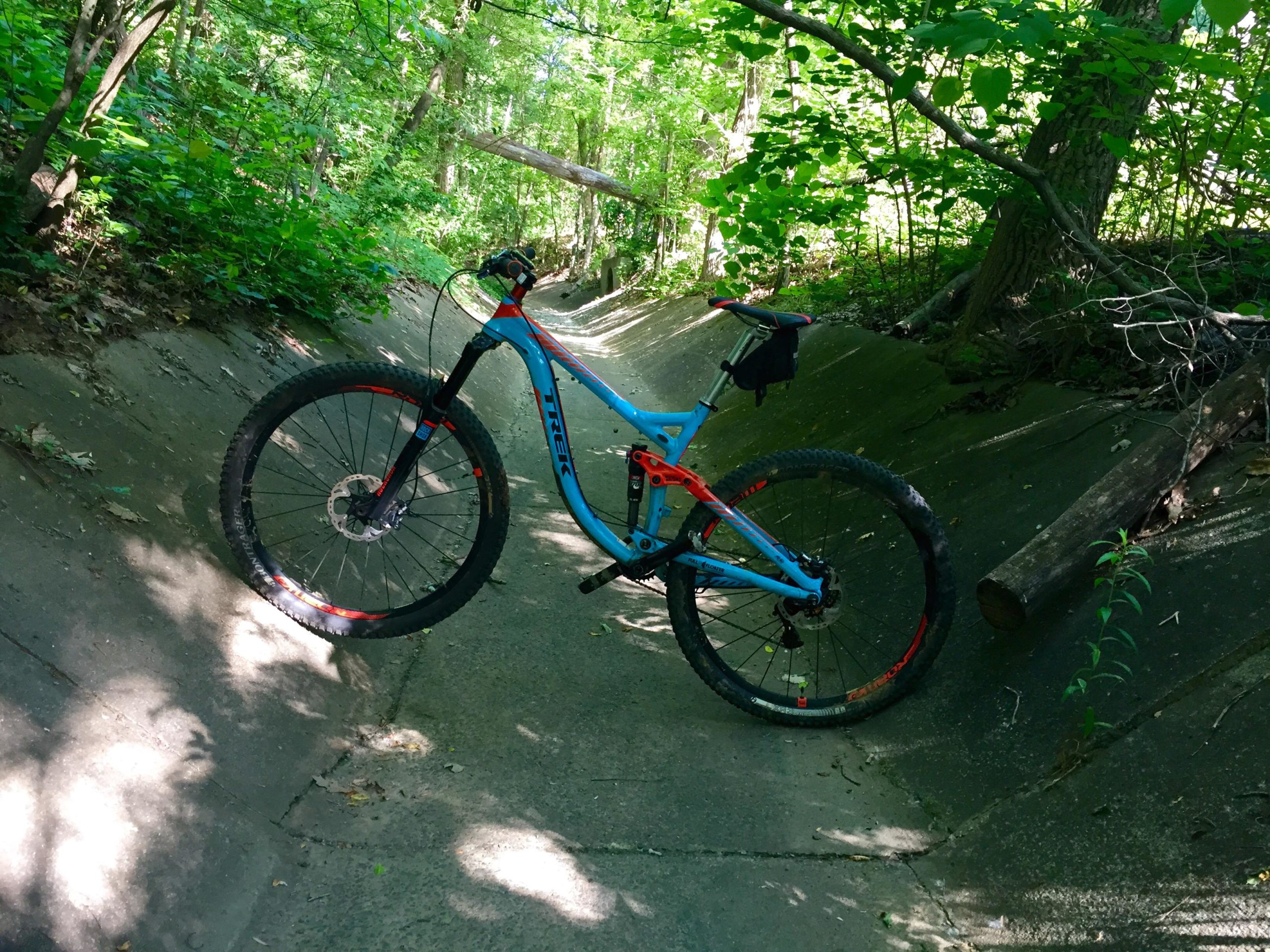 A blue and orange mountain bike is leaning against a concrete ditch surrounded by dense green foliage. Sunlight filters through the trees, casting dappled shadows on the ground. A fallen log lies nearby, adding to the natural setting. Back Yard Trails mountain bike trail.