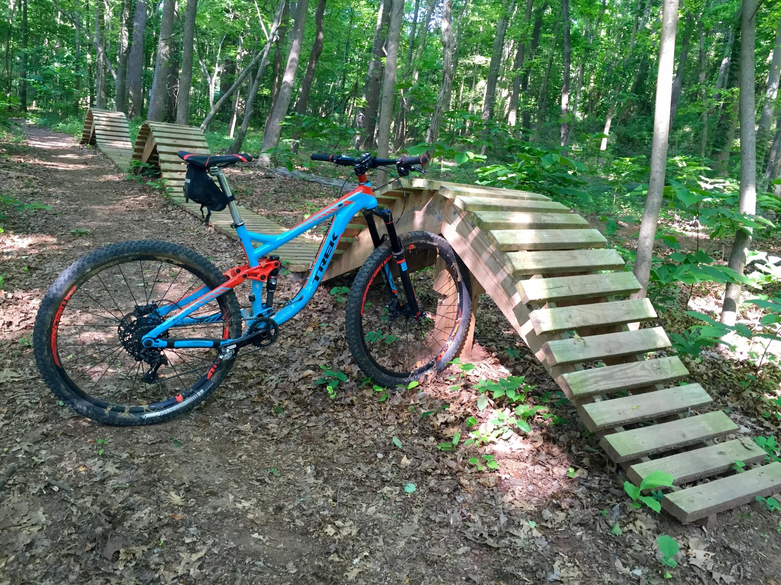 A blue and orange mountain bike is parked on a dirt trail in a wooded area, beside a wooden curved ramp designed for biking. The trail is surrounded by lush green foliage and trees, providing a natural setting for outdoor biking activities. Back Yard Trails mountain bike trail.