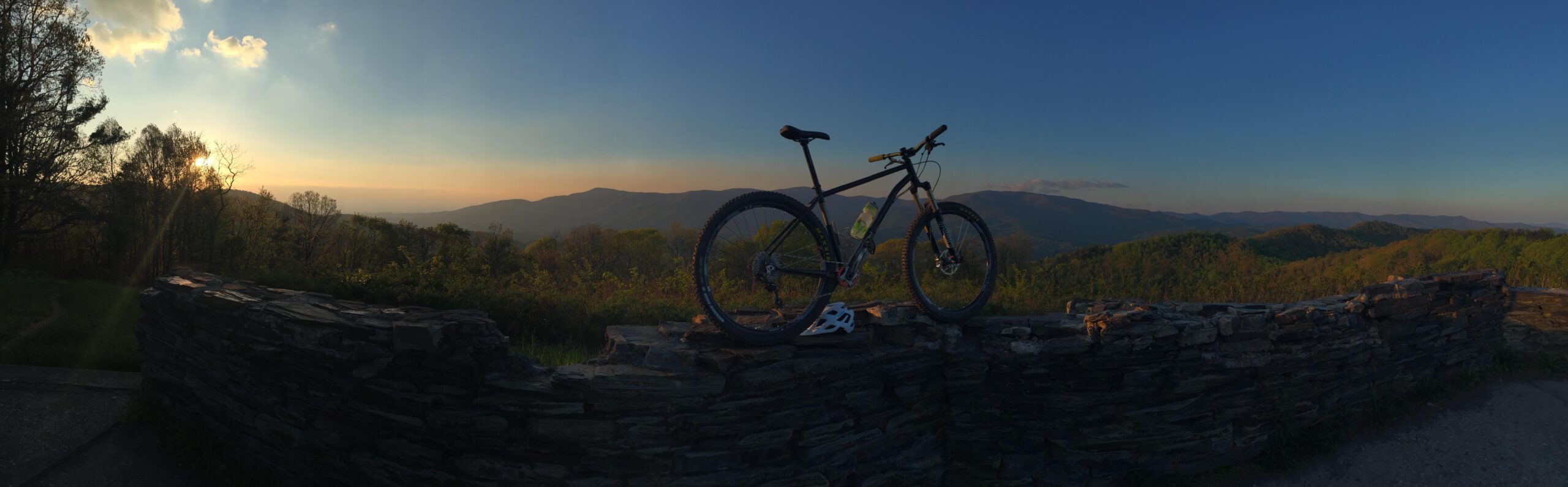 A mountain bike rests on a stone wall at sunset, overlooking a panoramic view of rolling hills and trees. The sky is filled with soft clouds and a warm glow as the sun sets in the distance, creating a serene outdoor scene. Pinhoti Trail: P3 mountain bike trail.