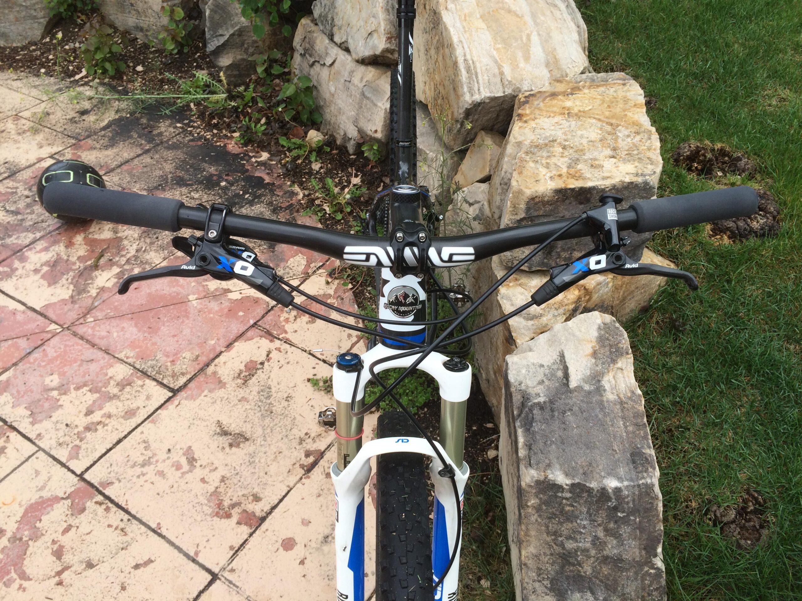 Rocky Mountain Vertex 990 rsl 2013: Alt text: Overhead view of mountain bike handlebars with grips and brake levers, partially resting on a stone wall with grass and paved surface in the background.
