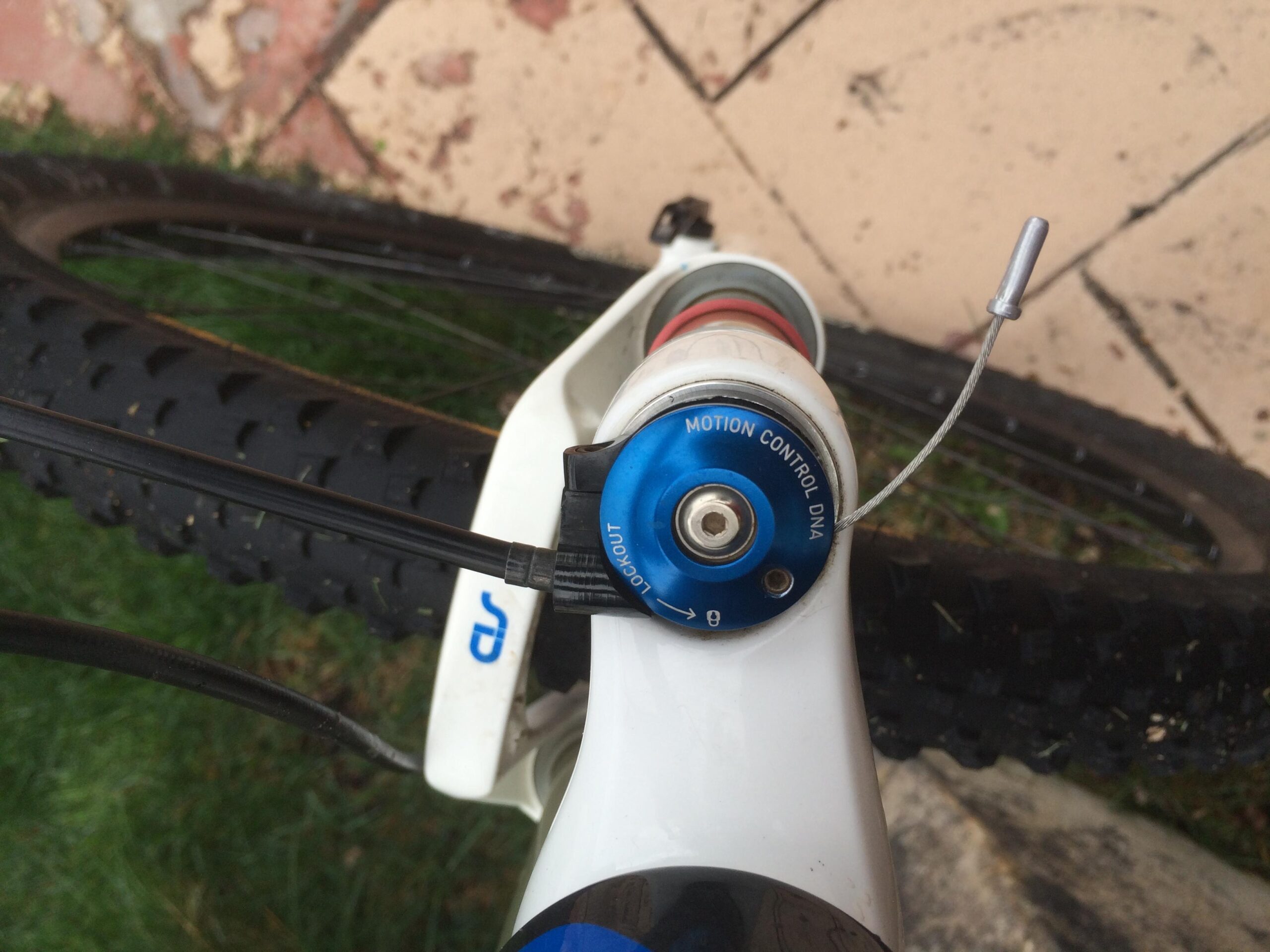 Rocky Mountain Vertex 990 rsl 2013: A close-up view of a mountain bike's suspension system, featuring a blue adjustment knob labeled "MOTION CONTROL DNA" and an attached cable running alongside a black tire. The background includes green grass and a tiled surface.