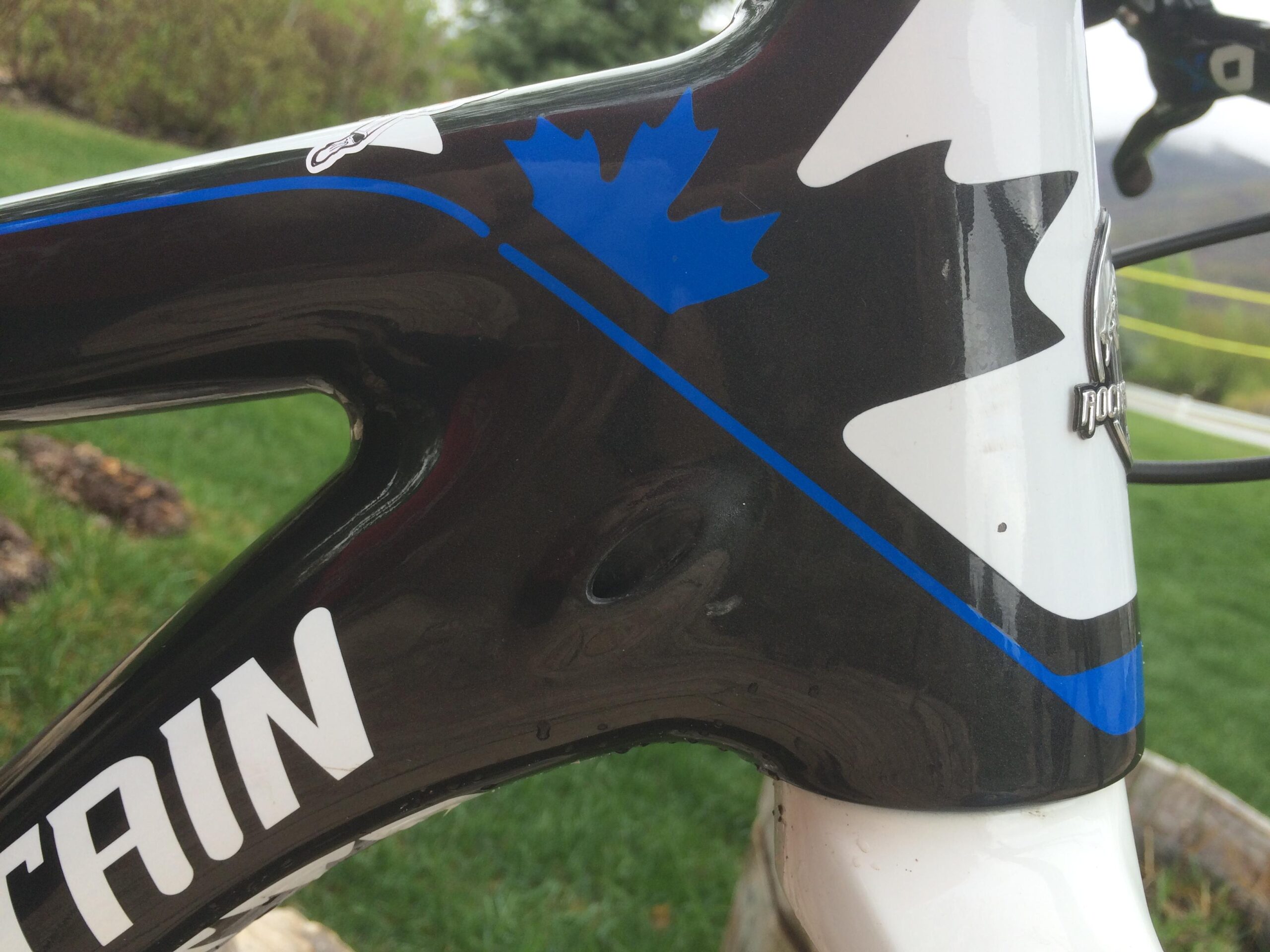 Rocky Mountain Vertex 990 rsl 2013: Close-up of a bicycle frame showing the head tube area, featuring a black and white design with blue and Canadian flag accents. The image captures the frame’s details against a backdrop of green grass and blurred landscape.